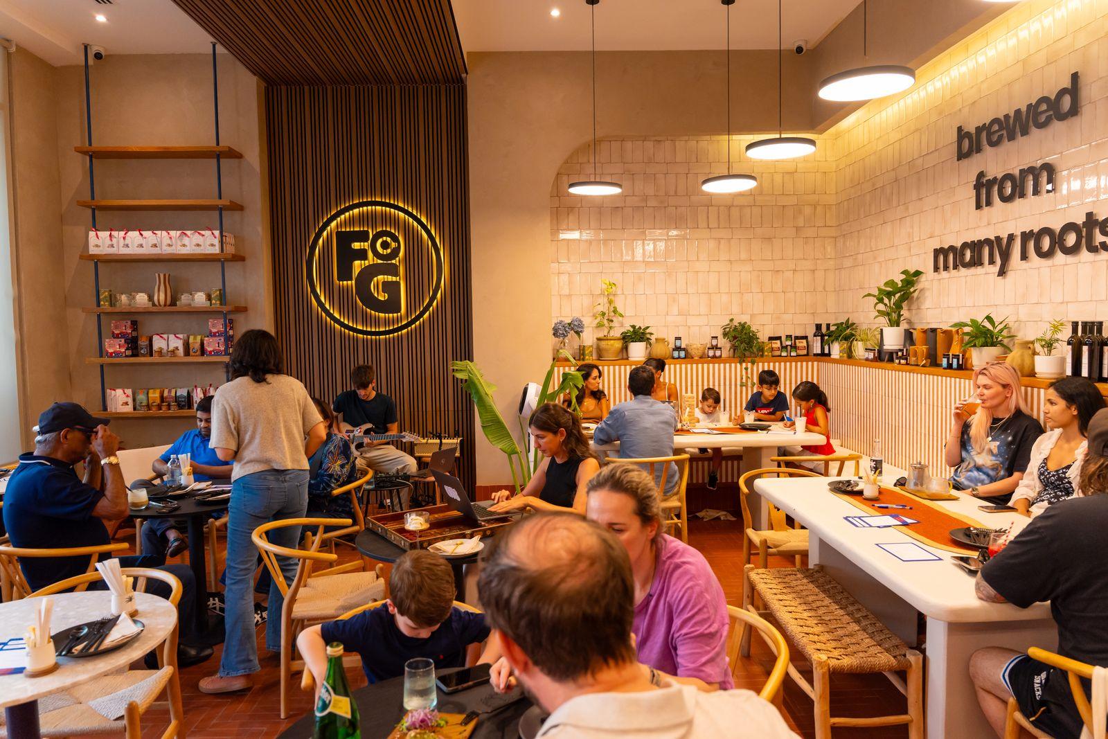 A full room of guests at Freshly Ground, with the illuminated FG logo and the 'brewed from many roots' wall behind them