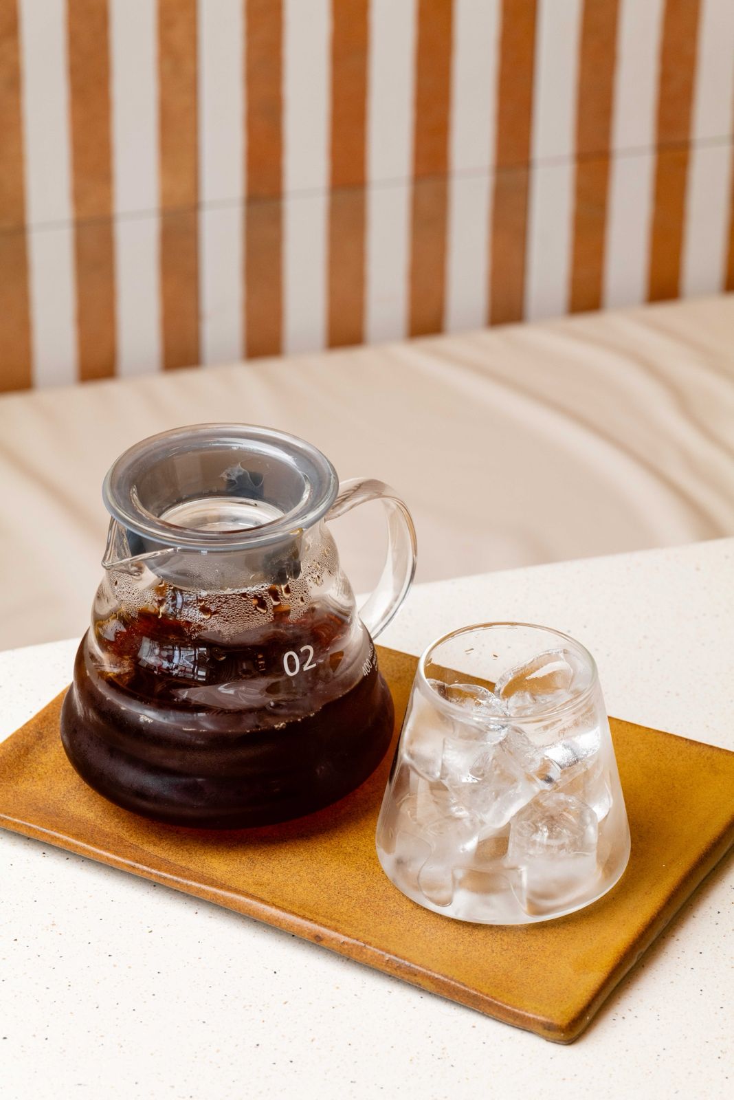 A glass carafe of cold brew coffee beside a tumbler of ice on a mustard ceramic tray at Freshly Ground