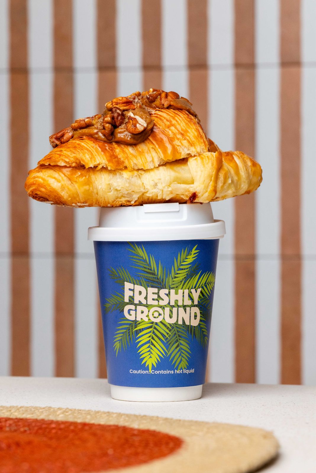 A pecan and caramel croissant balanced on top of a branded Freshly Ground takeaway cup
